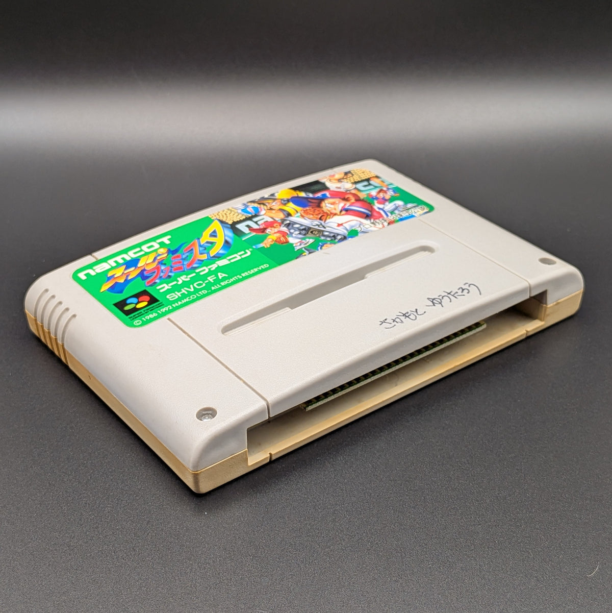 Super Famista video game cartridge for Nintendo's Super Famicom (SFC) Entertainment System, shown laying on its back with the label facing upwards. Showing the left hand side and bottom slot. It is displayed on a dark grey background.