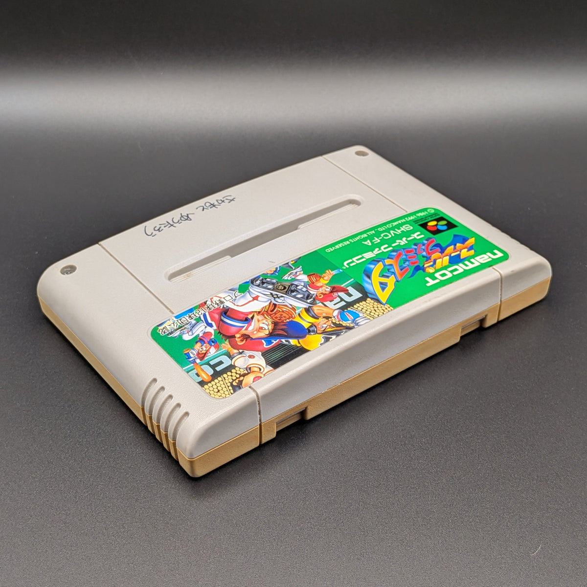 Super Famista video game cartridge for Nintendo's Super Famicom (SFC) Entertainment System, shown laying on its back with the label facing upwards. Showing the right hand side and top edge. It is displayed on a dark grey background.