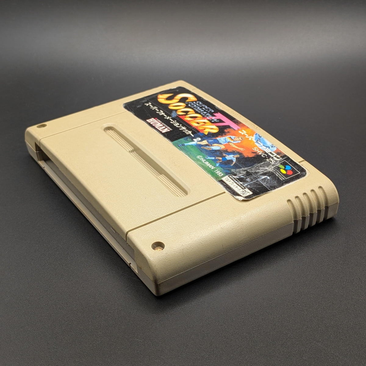 Super Formation Soccer 2 video game cartridge for Nintendo's Super Famicom (SFC) Entertainment System, shown laying down  on its back, on a dark grey background.
