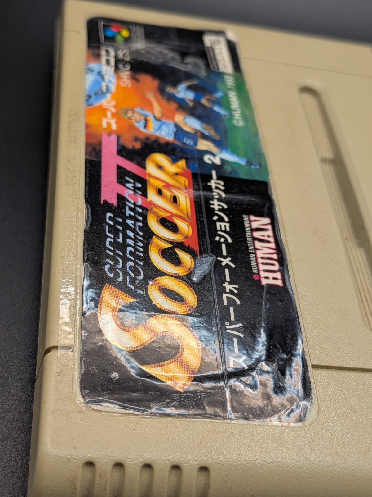 Super Formation Soccer 2 video game cartridge for Nintendo's Super Famicom (SFC) Entertainment System, showing a close up of the tapped label, on a dark grey background.
