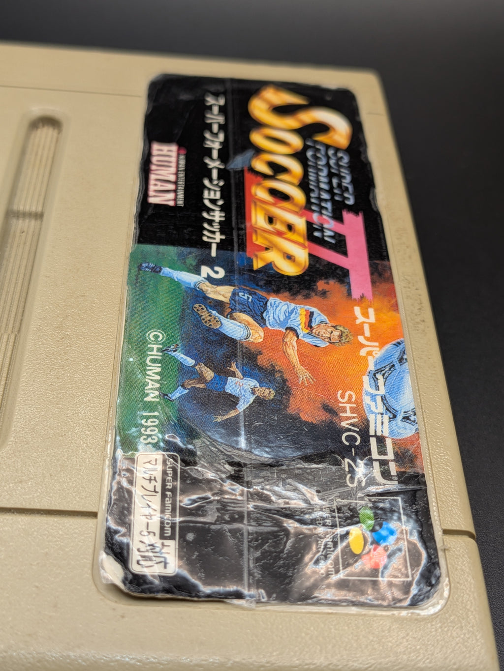 Super Formation Soccer 2 video game cartridge for Nintendo's Super Famicom (SFC) Entertainment System, showing a close up of the tapped label, on a dark grey background.
