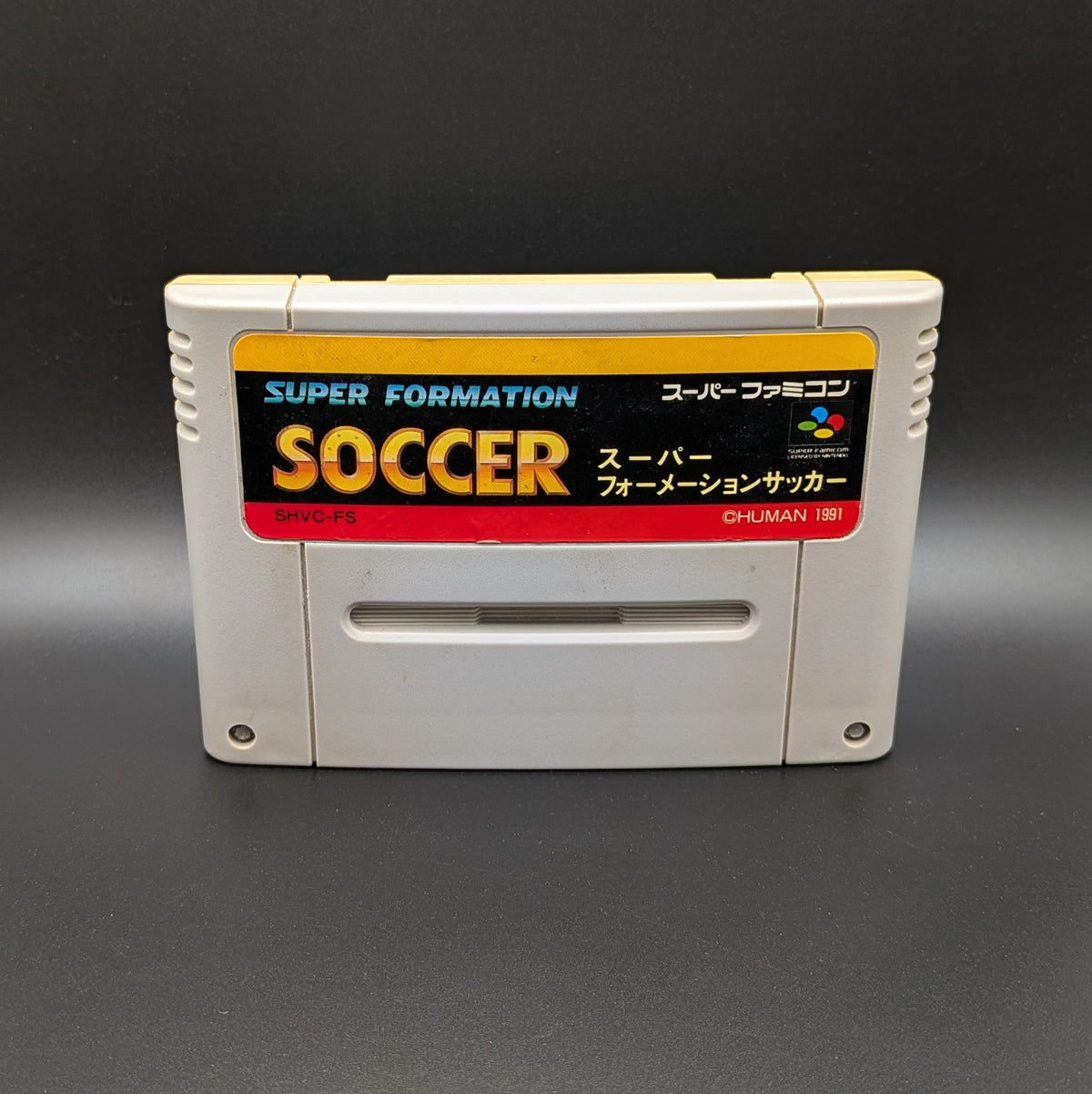 Super Formation Soccer video game cartridge for Nintendo's Super Famicom (SFC) Entertainment System, shown standing up and facing forwards on a dark grey background.