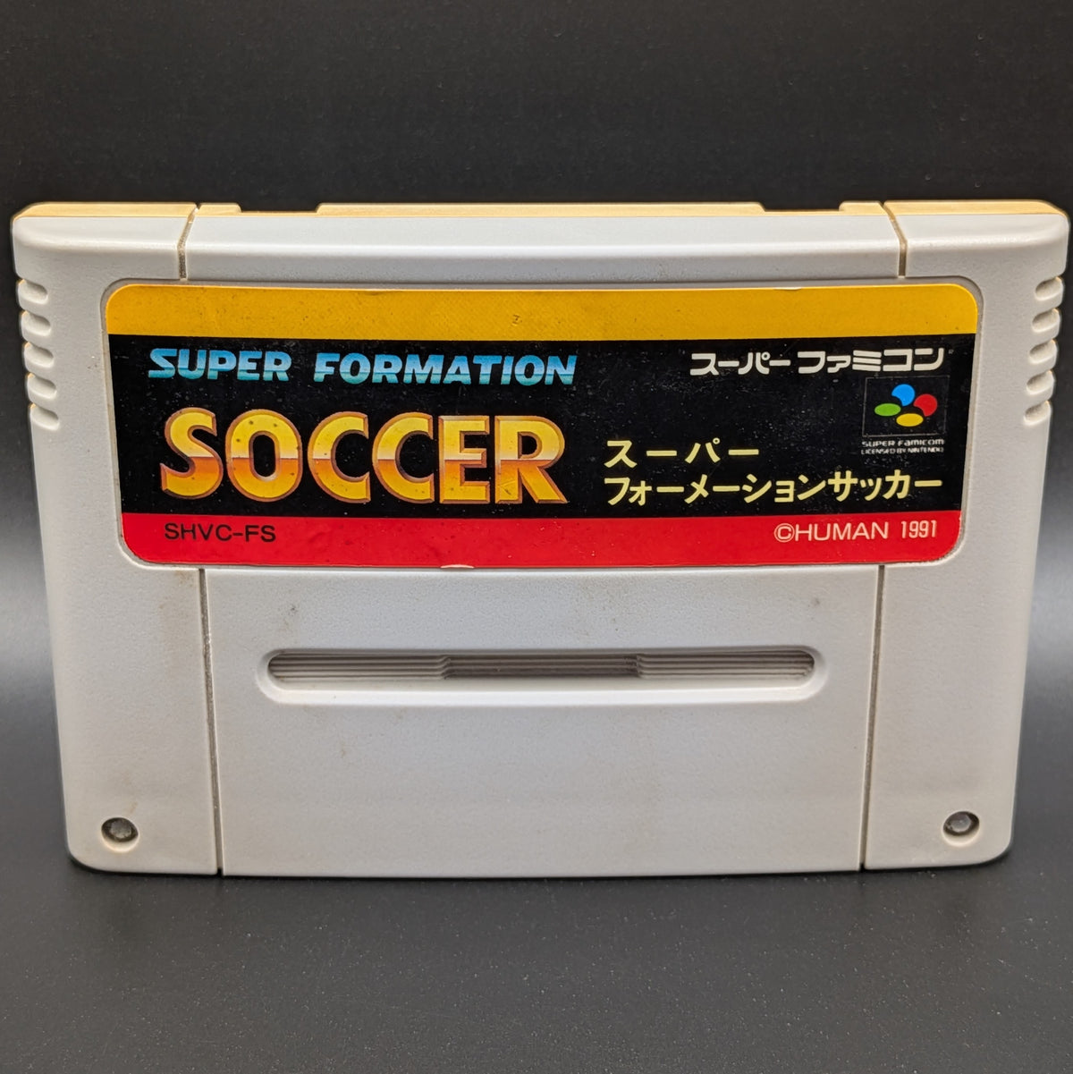 A close up of Super Formation Soccer video game cartridge for Nintendo's Super Famicom (SFC) Entertainment System, shown standing up and facing forwards on a dark grey background.