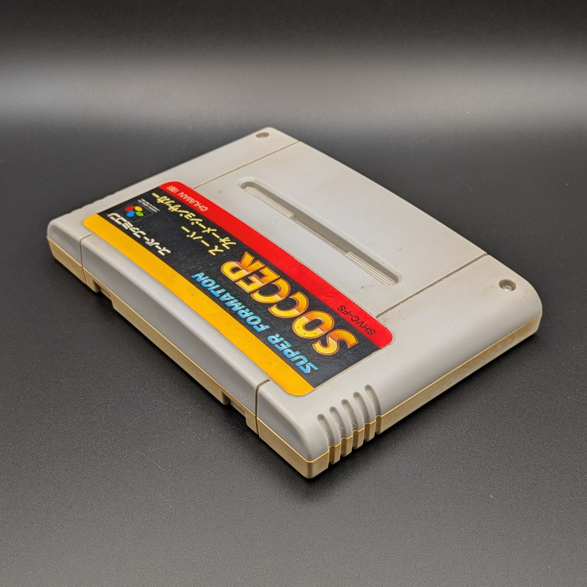 Super Formation Soccer video game cartridge for Nintendo's Super Famicom (SFC) Entertainment System, shown laying on its back with the label facing upwards. Showing the left hand side and top edge. It is displayed on a dark grey background.
