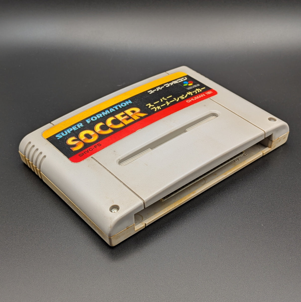 Super Formation Soccer video game cartridge for Nintendo's Super Famicom (SFC) Entertainment System, shown laying on its back with the label facing upwards. Showing the left hand side and bottom slot. It is displayed on a dark grey background.