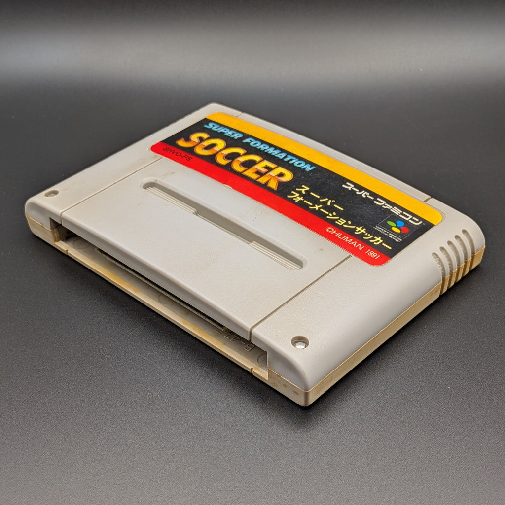 Super Formation Soccer video game cartridge for Nintendo's Super Famicom (SFC) Entertainment System, shown laying on its back with the label facing upwards. Showing the right hand side and bottom slot. It is displayed on a dark grey background.