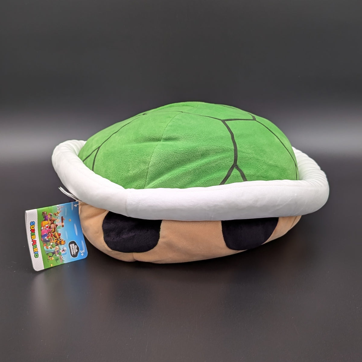 A plush Super Mario turtle shell pillow with a green shell, white trim and beige base, shown on a dark grey background with a product tag attached.
