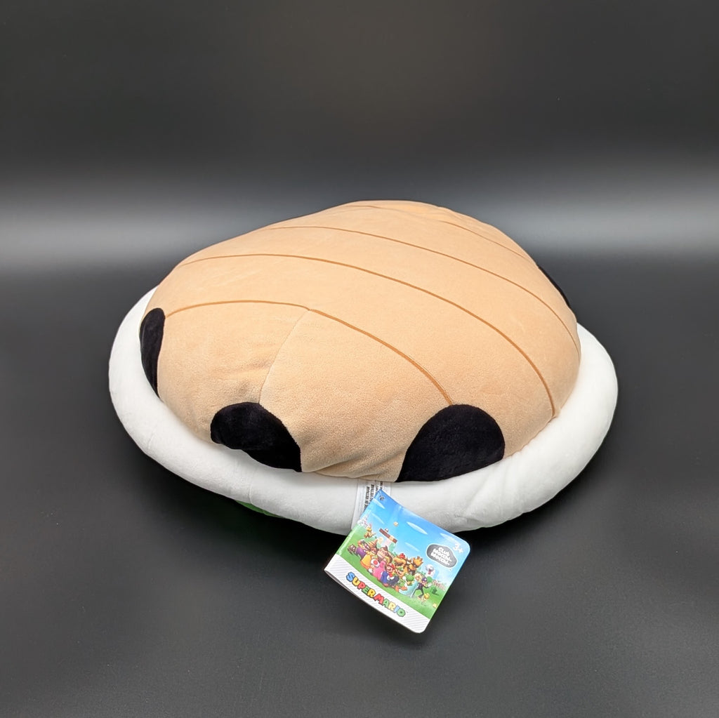 A  mega green shell plush with a white padded rim, laying on its back to show the tan underside and with its Super Mario tag visible, photographed on a dark grey background. From Nintendo's beloved Super Mario and Mario Kart games,