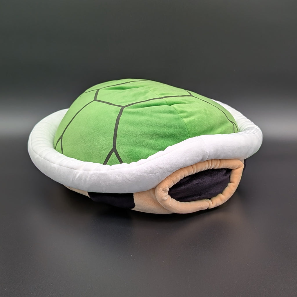 A green turtle shell plushie with a white padded rim, laying on its back to show the tan underside, and black accent details, photographed on a dark grey background. From Nintendo's beloved Super Mario and Mario Kart games,