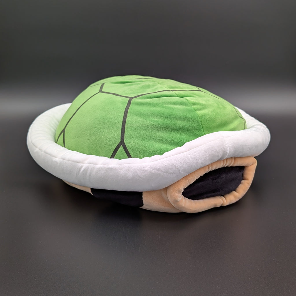 A plush Super Mario green shell plush pillow, with white trim and beige base, shown on a dark grey background with a product tag attached. From Nintendo's beloved Super Mario and Mario Kart games,