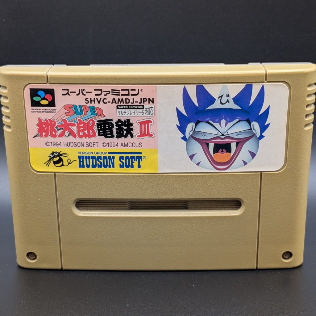 A close up of Super Momotarou Dentetsu 3 video game cartridge for Nintendo's Super Famicom (SFC) Entertainment System, shown standing up and facing forwards on a dark grey background.