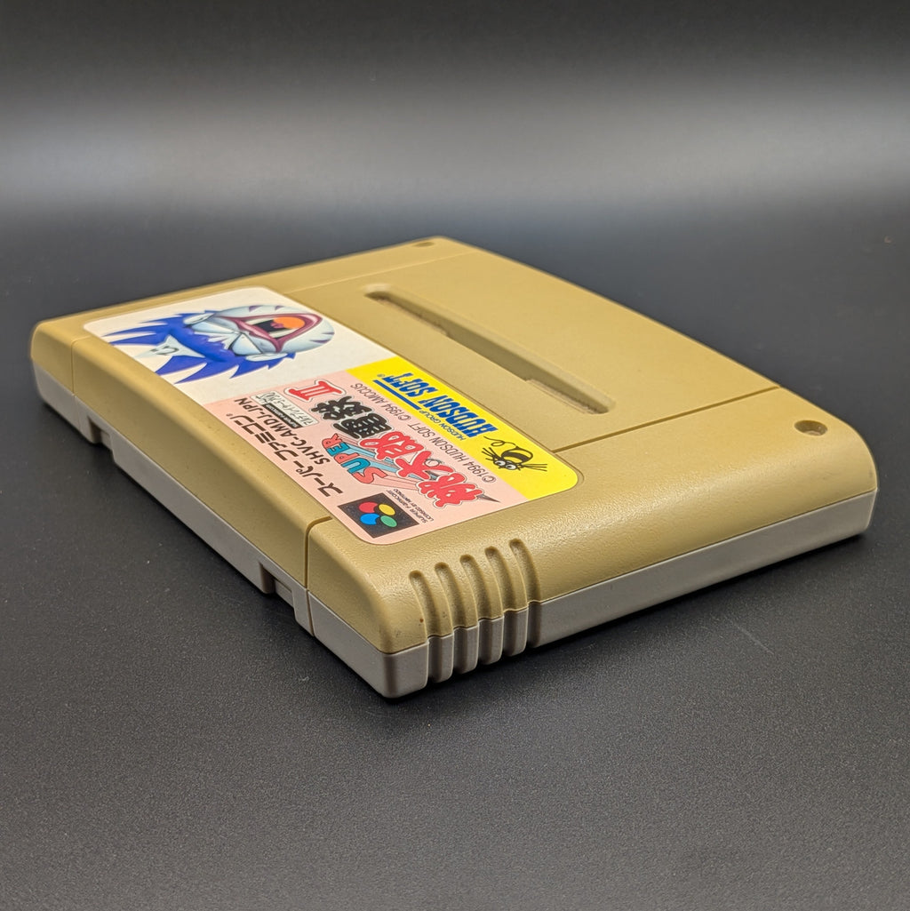 Super Momotarou Dentetsu 3 video game cartridge for Nintendo's Super Famicom (SFC) Entertainment System, shown laying on its back with the label facing upwards. Showing the left hand  and top edge. It sits on a dark grey background