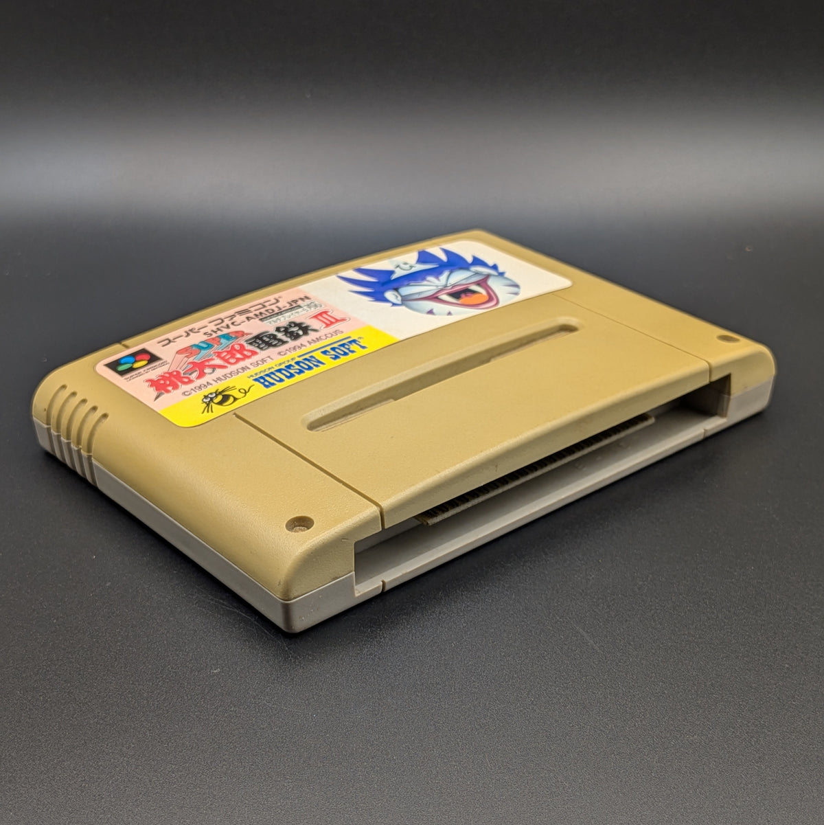 Super Momotarou Dentetsu 3 video game cartridge for Nintendo's Super Famicom (SFC) Entertainment System, shown laying on its back with the label facing upwards. Showing the left hand side and bottom slot. It sits on a dark grey background