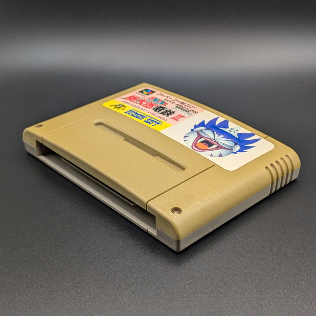 Super Momotarou Dentetsu 3 video game cartridge for Nintendo's Super Famicom (SFC) Entertainment System, shown laying on its back with the label facing upwards. Showing the right hand side and bottom slot. It sits on a dark grey background.