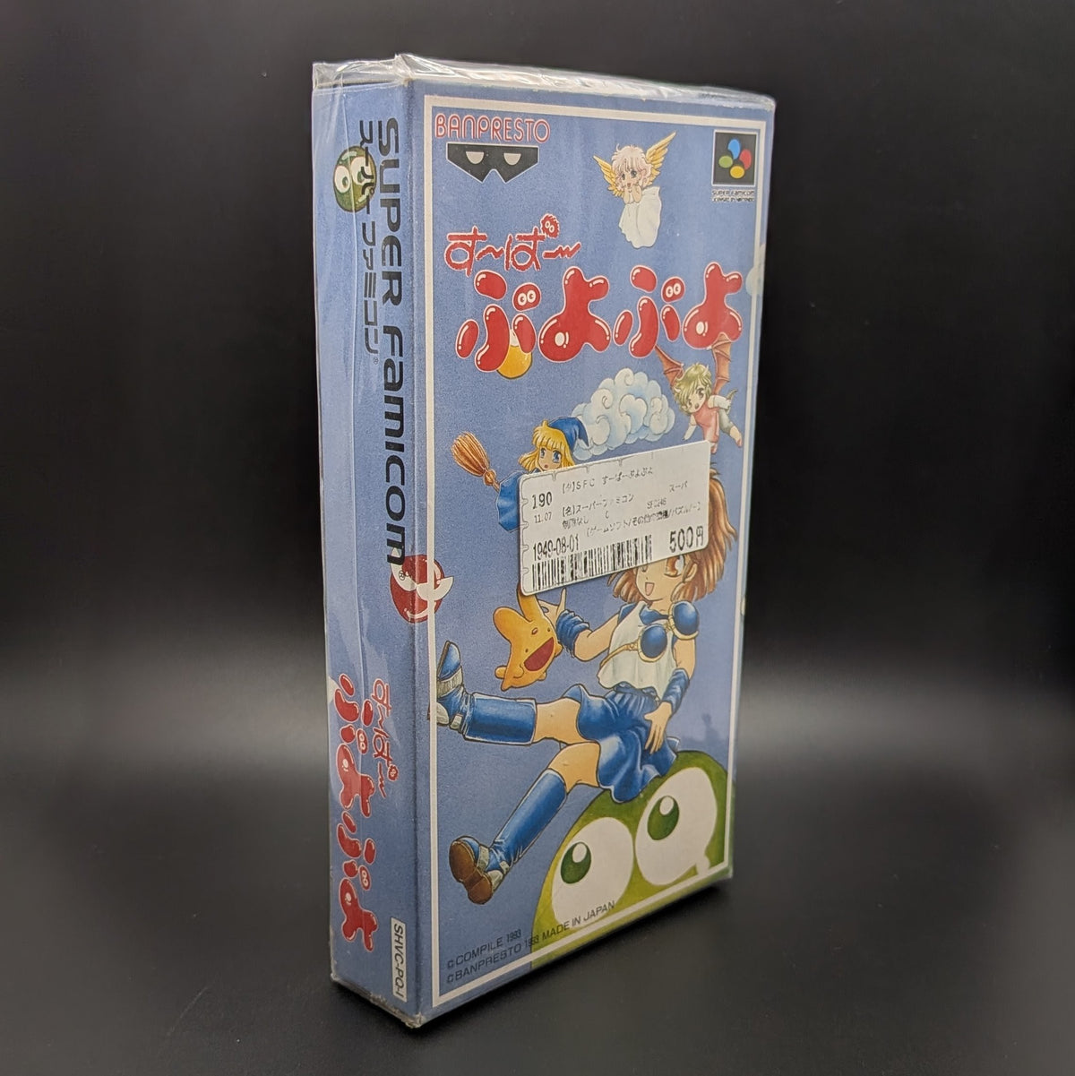 Super Puyo Puyo boxed and sealed video game cartridge for Nintendo's Super Famicom (SFC) Entertainment System, shown standing up at 3/4 angle facing the right, central on a dark grey background. There is a Japanese store sticker stuck to the front on the outer packaging.
