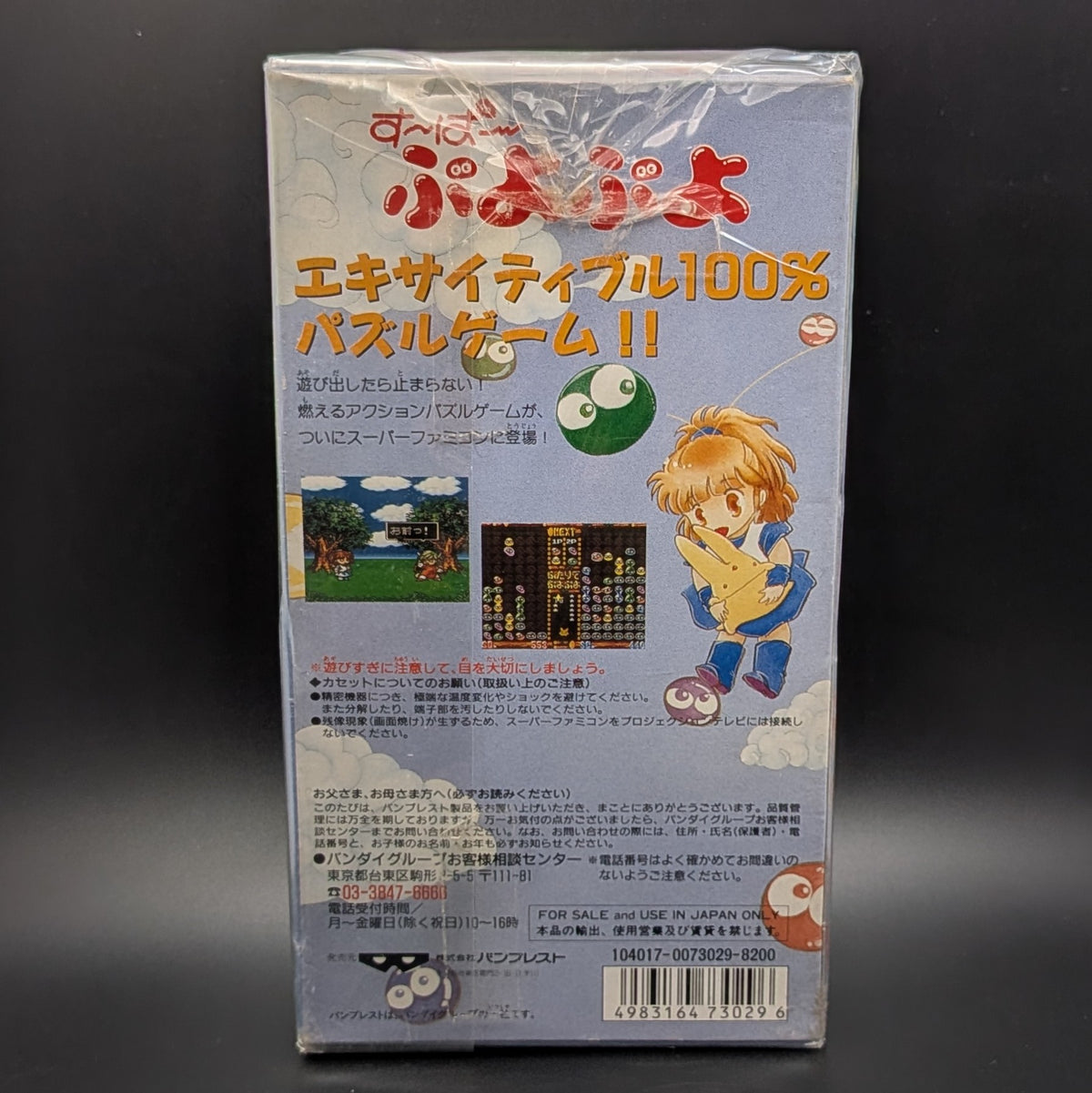 Super Puyo Puyo boxed and sealed video game cartridge for Nintendo's Super Famicom (SFC) Entertainment System, shown standing up and showing the back, central on a dark grey background.The back of the box features a light blue background with Japanese text, a cartoon girl character, and small screenshots of the game.