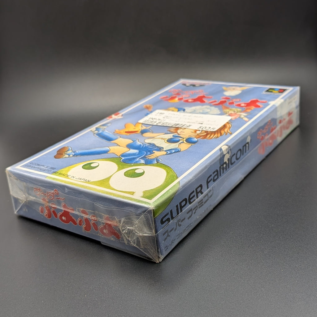 Super Puyo Puyo boxed and sealed video game cartridge for Nintendo's Super Famicom (SFC) Entertainment System, shown laying on it back at 3/4 angle, on a dark grey background. There is a Japanese store sticker stuck to the front on the outer packaging.