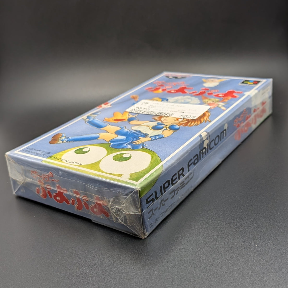 Super Puyo Puyo boxed and sealed video game cartridge for Nintendo's Super Famicom (SFC) Entertainment System, shown laying on it back at 3/4 angle, on a dark grey background. There is a Japanese store sticker stuck to the front on the outer packaging.