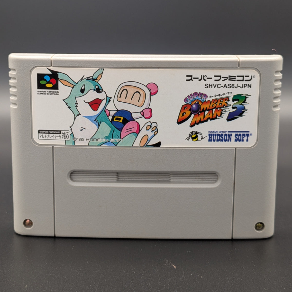 A close up of Super Bomberman 3 video game cartridge for Nintendo's Super Famicom (SFC) Entertainment System, shown standing up and facing forwards on a dark grey background.