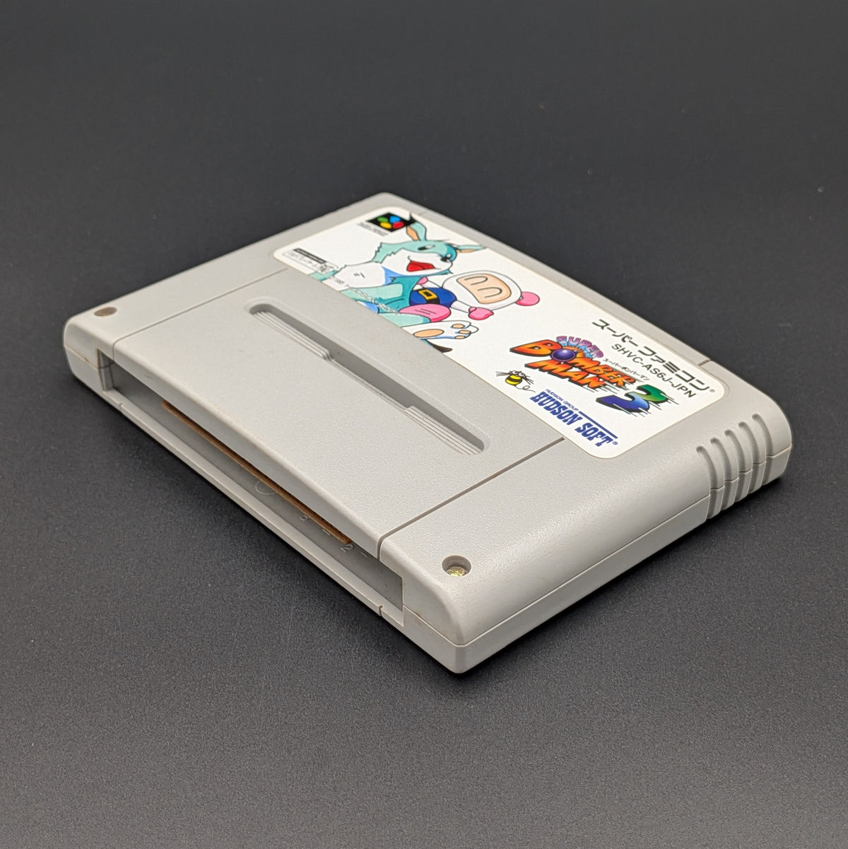 Super Bomberman 3 video game cartridge for Nintendo's Super Famicom (SFC) Entertainment System, shown laying on its back with the label facing upwards. Showing the right hand side and bottom slot. It is displayed on a dark grey background.