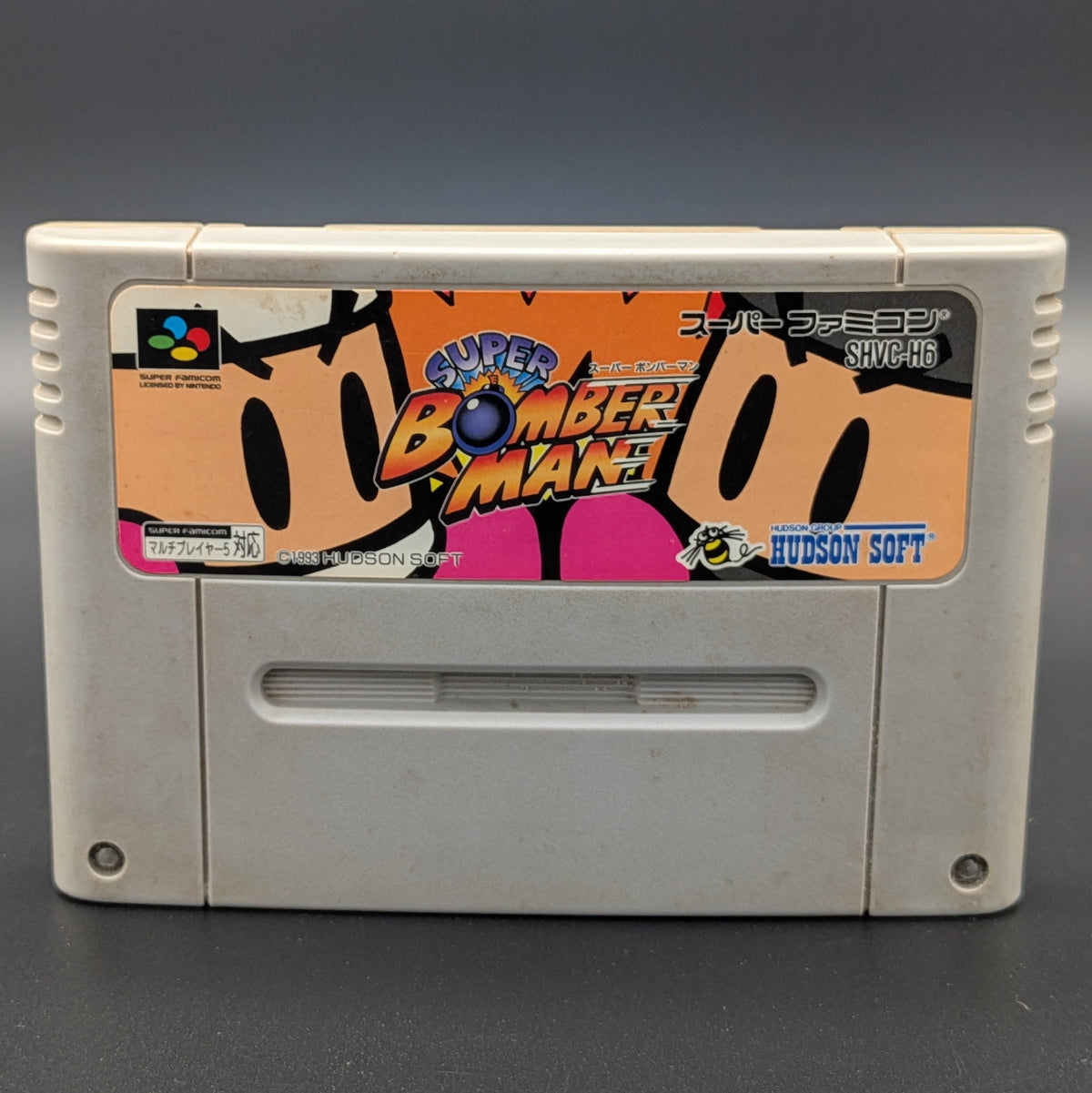 A close up of Super Bomberman video game cartridge for Nintendo's Super Famicom (SFC) Entertainment System, shown standing up and facing forwards on a dark grey background.