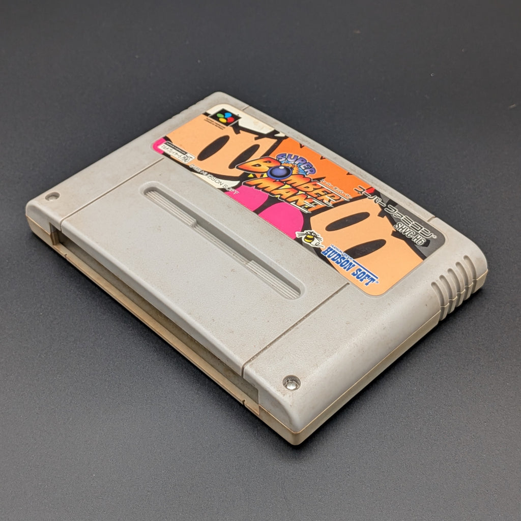 Super Bomberman video game cartridge for Nintendo's Super Famicom (SFC) Entertainment System, shown laying on its back with the label facing upwards. Showing the right hand side and bottom slot. It is displayed on a dark grey background.