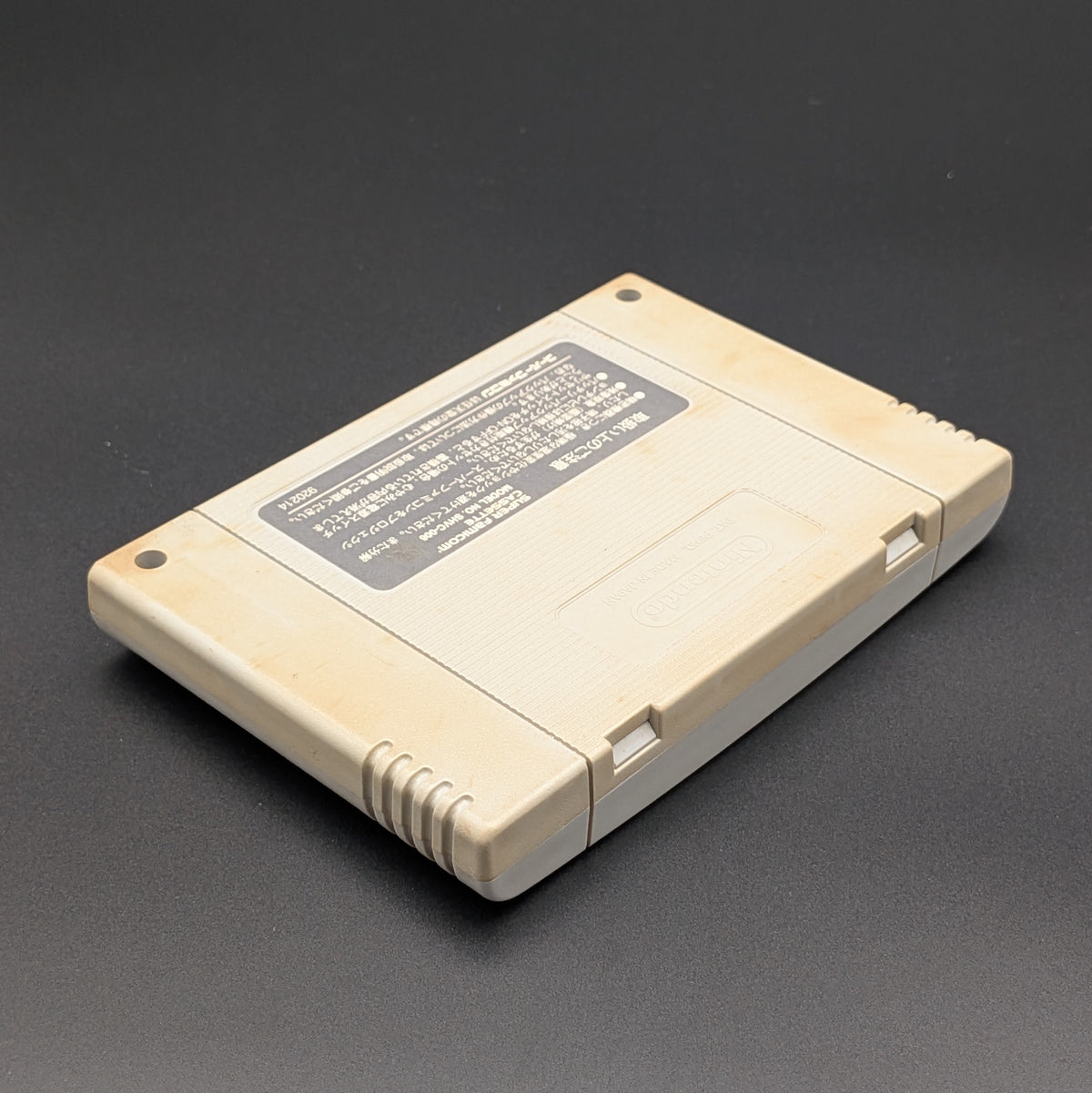 Super Bomberman video game cartridge for Nintendo's Super Famicom (SFC) Entertainment System, shown laying on its face with the back label facing upwards. Showing the right hand side and top edge. It is displayed on a dark grey background.