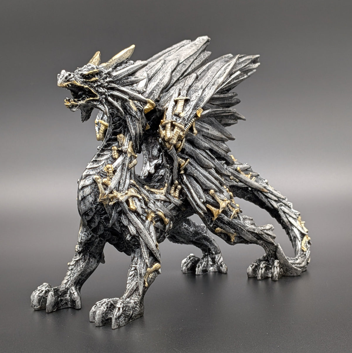 A detailed fantasy dragon figurine in a metallic silver-gray finish with gold accents, standing on a dark gray surface against a matching gray gradient background.