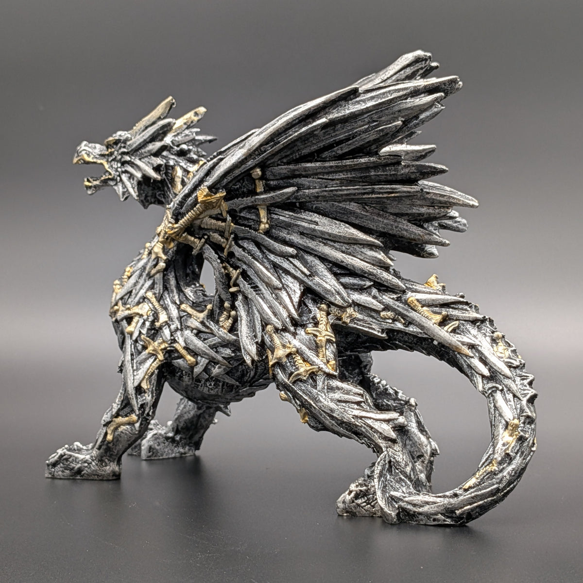 A detailed fantasy dragon figurine crafted from metallic silver swords with small gold accents, shown in a dynamic crouching pose against a dark gray gradient background.