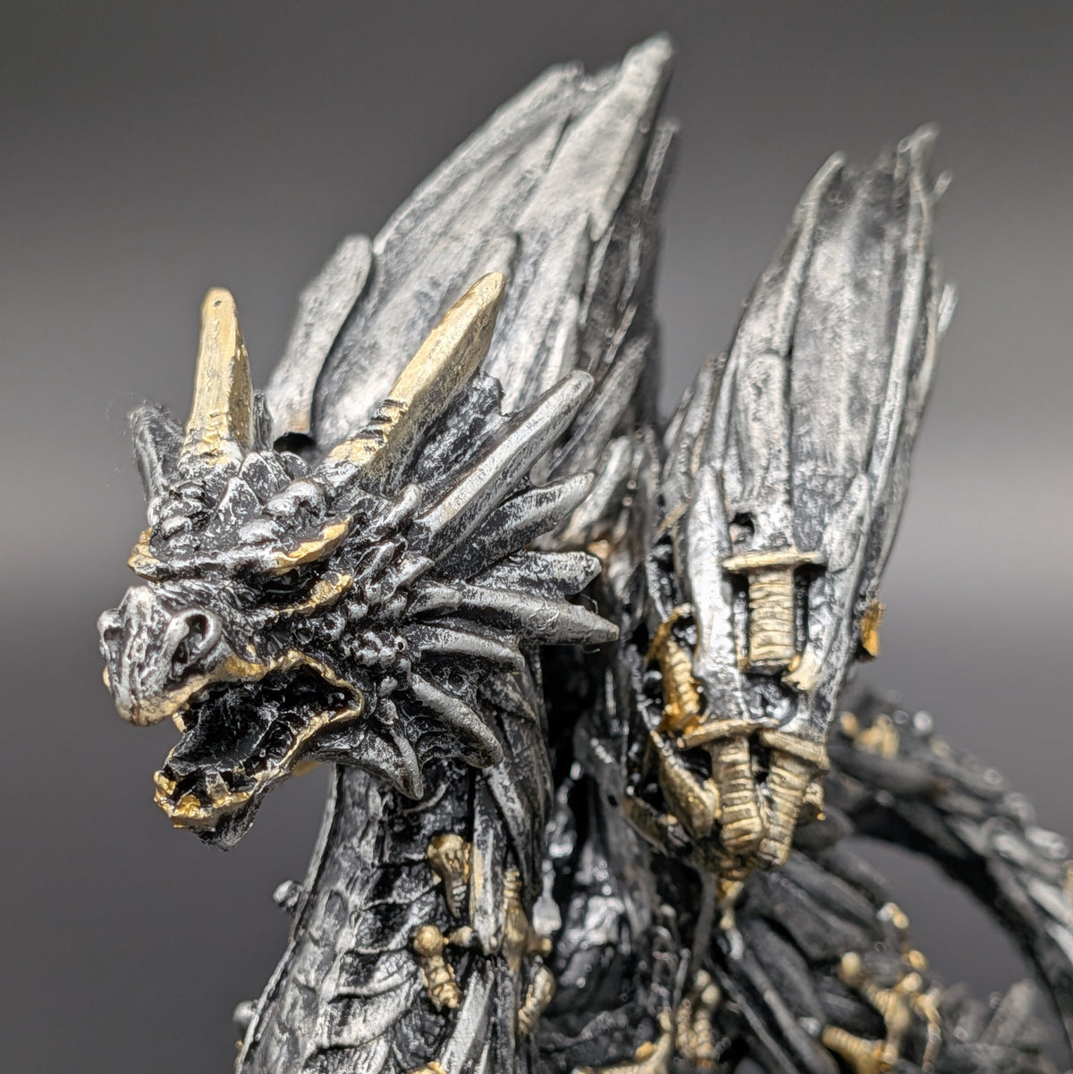 Close-up of a detailed fantasy dragon figurine with metallic silver-gray scales and gold accents on its horns, teeth, and skeletal details, set against a dark gray gradient background.