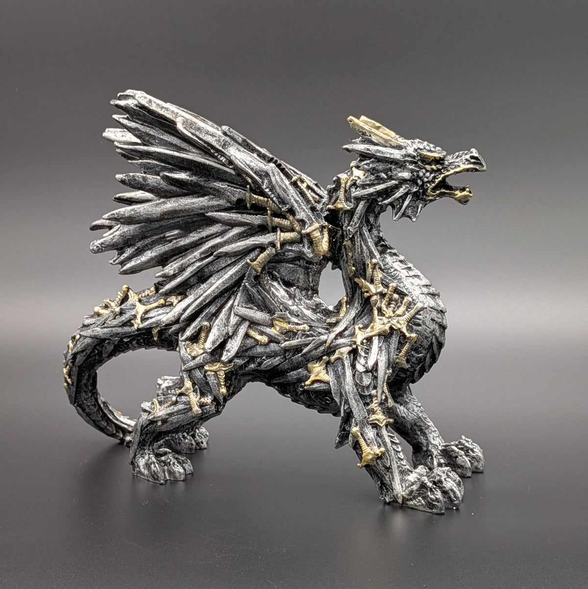 A detailed fantasy dragon figurine sculpted from metallic swords with gold accents, standing on a dark gray gradient background.