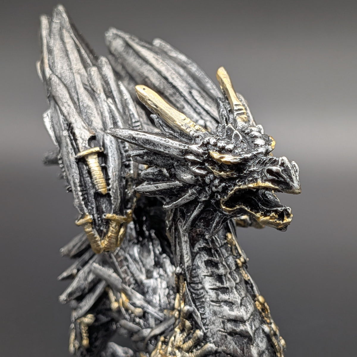 A close-up of a detailed fantasy dragon figurine with textured black and silver scales and gold accents, shown against a dark gray gradient background.
