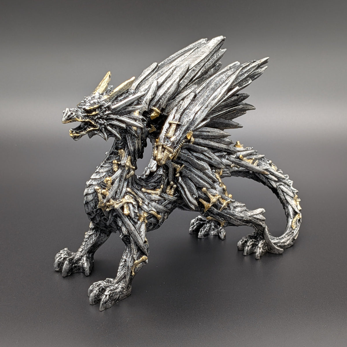 A highly detailed fantasy dragon figurine with textured scales, metallic silver-gray coloring, and small gold accents on its horns, claws, and armor, set against a dark gray gradient background.