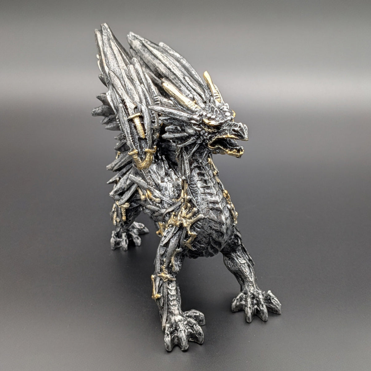 A detailed fantasy dragon figurine in a metallic silver and black textured finish with gold accents, standing on a dark gray gradient background.