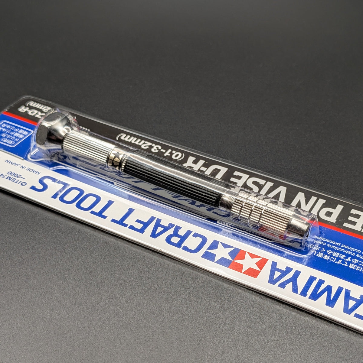 Tamiya 74112 Fine Pin Vise D-R (0.1-3.2mm) in its original blister card packaging, a close up of the tool through the clear packaging and displayed on a dark gray background.