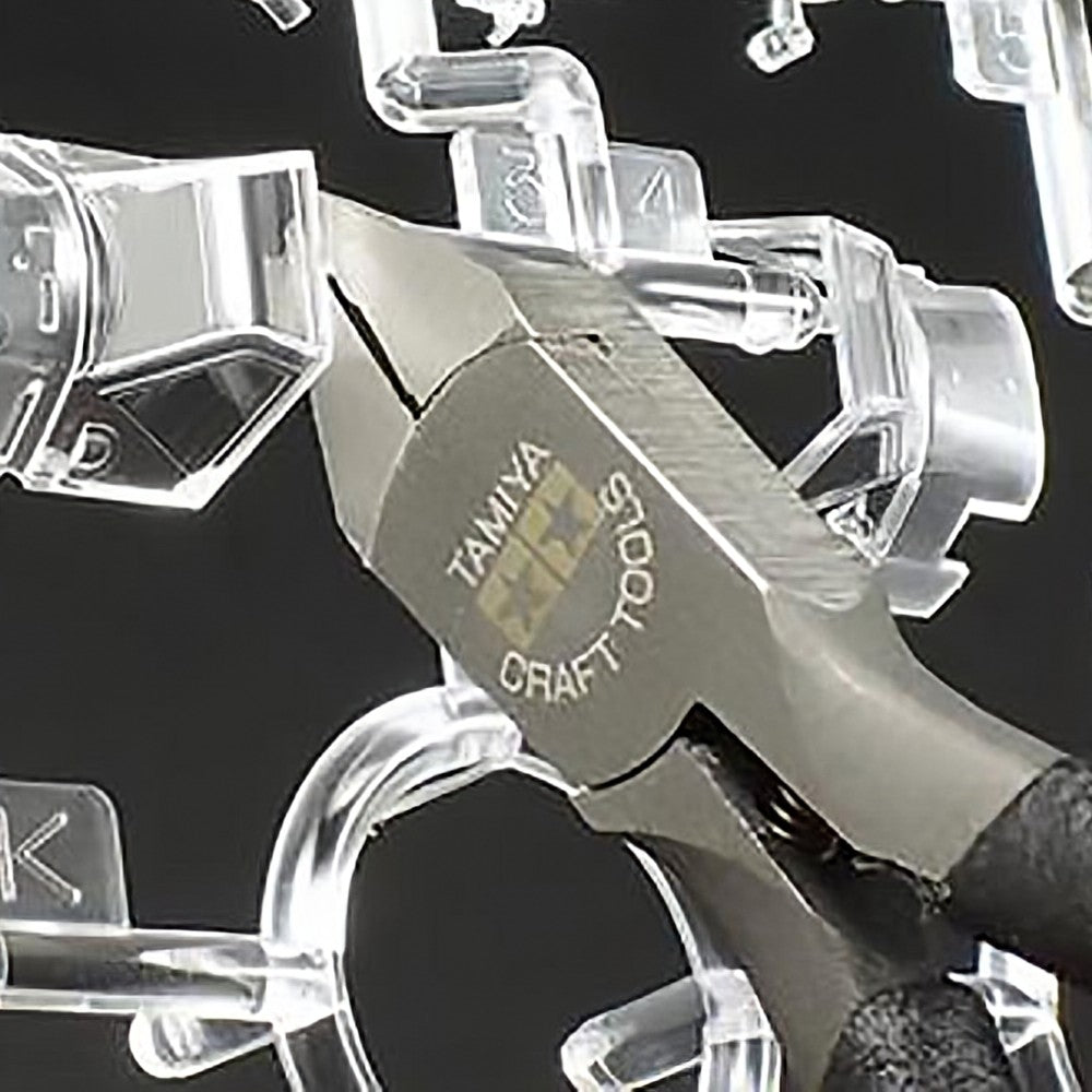 Close-up of a Tamiya craft tools Sharp Pointed Side Cutter, with a dark grey metal head and black handle, shown against a black background, snipping clear plastic pieces/sprues.