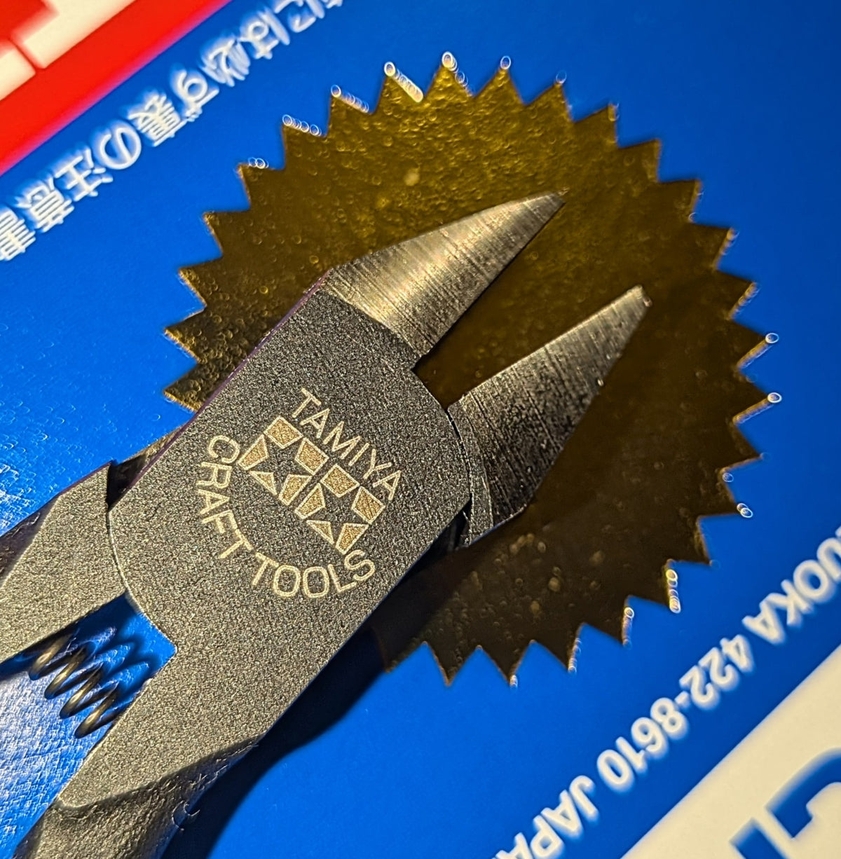 Close-up of a Tamiya Craft Tools sharp pointed side cutters for plastic in black, with a blue cardboard backing and gold starburst design behind the head of the cutters.