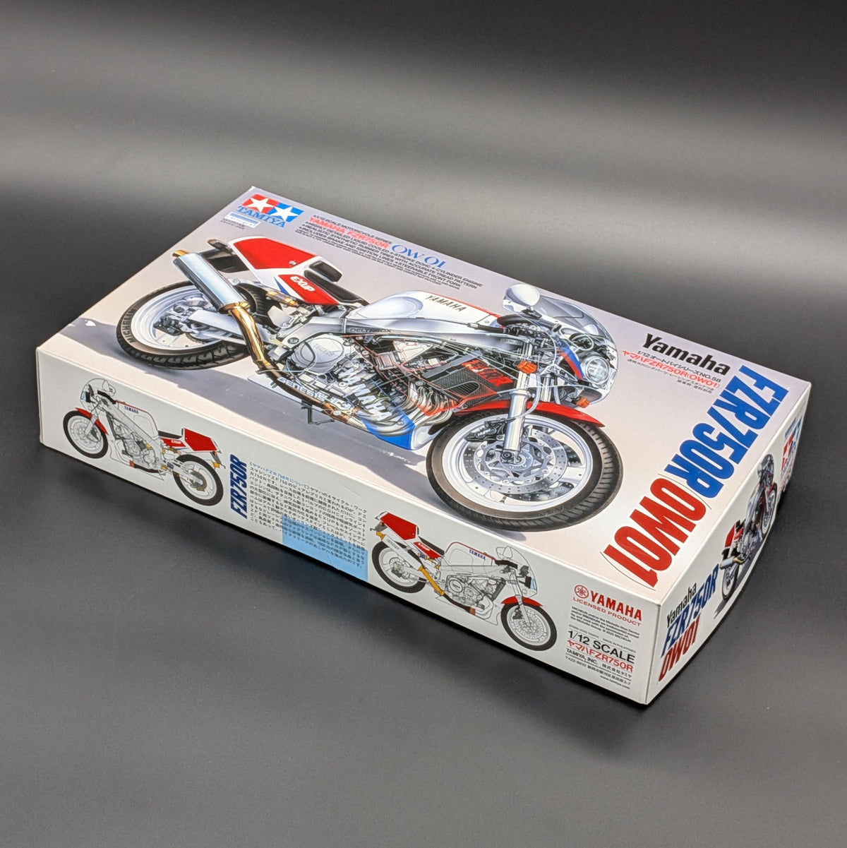 A boxed model kit of a Yamaha FZR750R OW01 motorcycle, shown at an angle on a dark gray gradient background. The packaging features a detailed illustration of the red, white, and blue motorcycle.