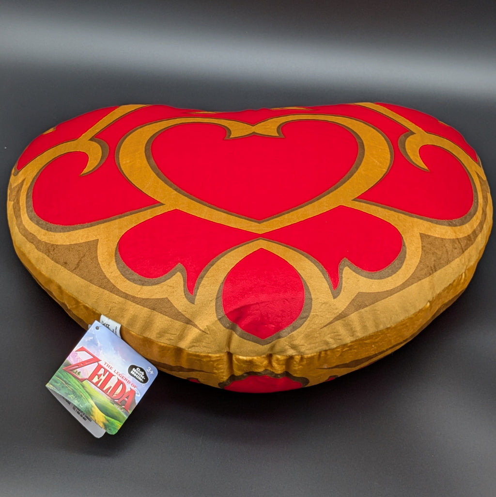 A heart-shaped plush pillow featuring a red and gold Zelda-themed design, with a visible 'The Legend of Zelda' tag attached, set against a dark gray background.