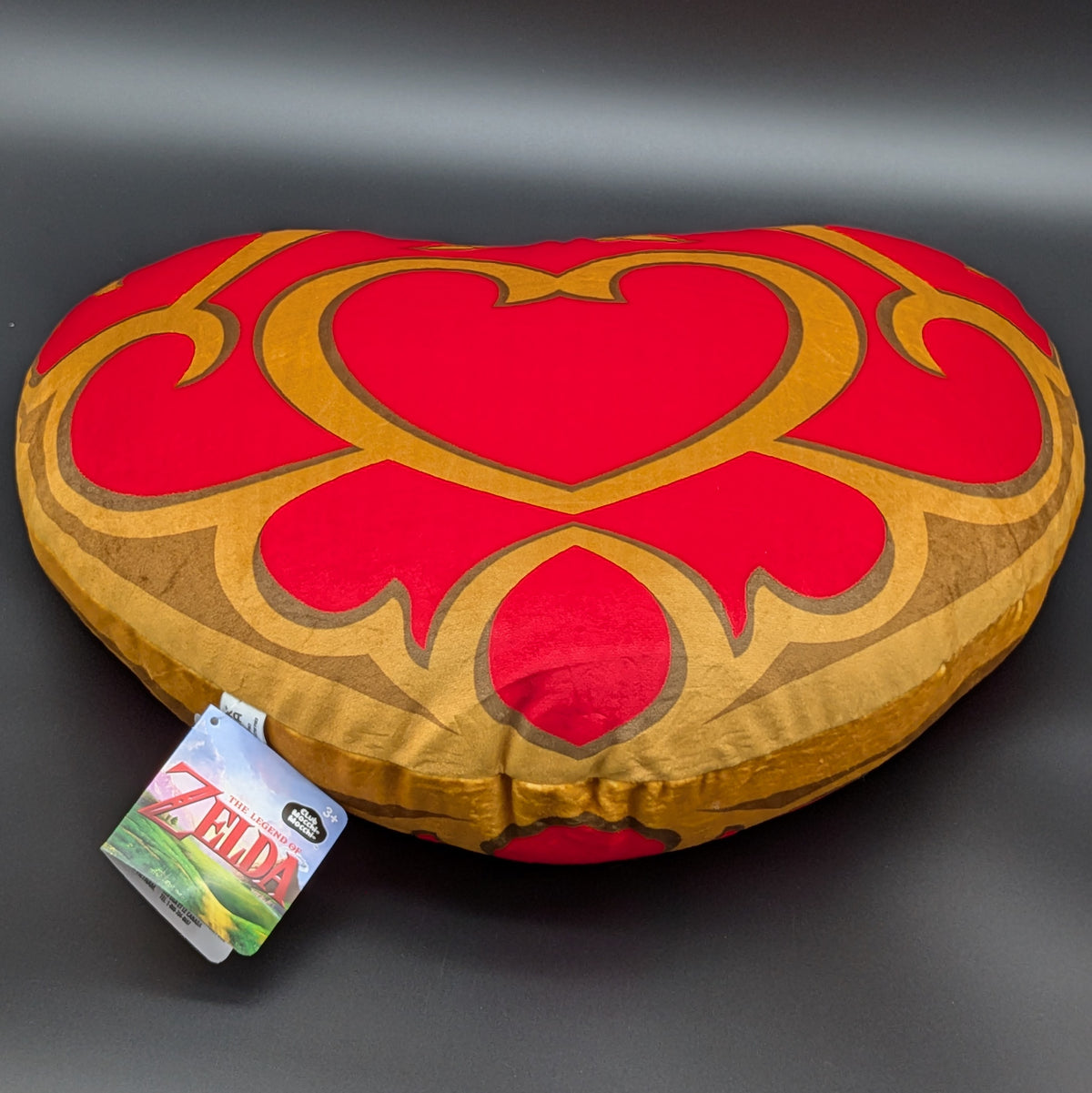 A heart-shaped plush pillow featuring a red and gold Zelda-themed design, with a visible 'The Legend of Zelda' tag attached, set against a dark gray background.