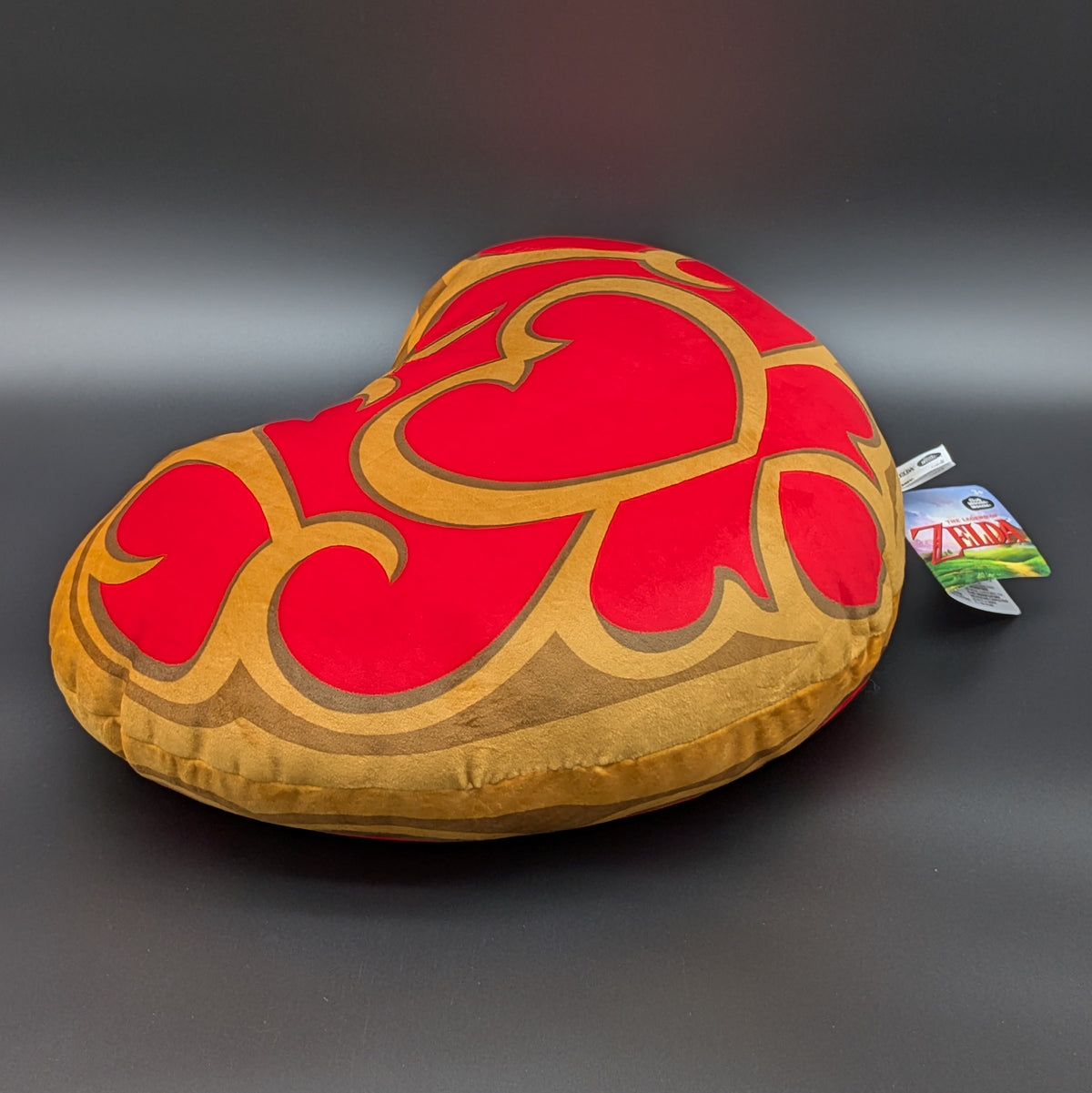 The Legend of Zelda, Link's Heart Container is a heart-shaped decorative plush/pillow with a red and gold pattern, shown laying down at a 3/4 view angle, on a dark grey background with its retail tag attached.