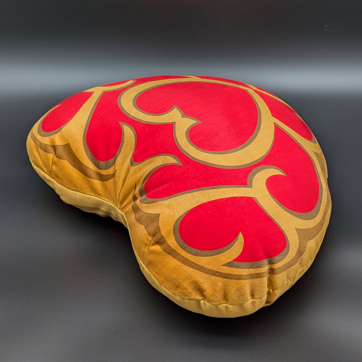 A heart-shaped decorative cushion with a red and gold scrollwork pattern, photographed on a dark grey background. This is a plushie of Links Heart Container, from The Legend of Zelda.