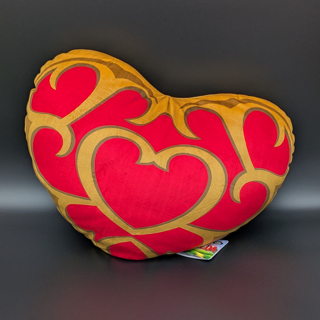 The Legend of Zelda, Link's Heart Container is a heart-shaped decorative plush/pillow with a red and gold pattern, shown face on & leaning to the right, on a dark grey background with its retail tag attached.