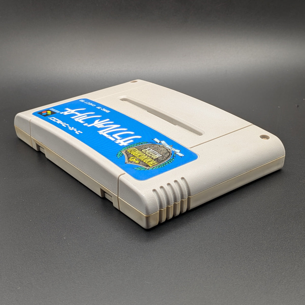 Thoroughbred Breeder video game cartridge for Nintendo's Super Famicom (SFC) Entertainment System, shown laying on its back with the label facing up and the top edge is visible. Pictured on a dark grey background.