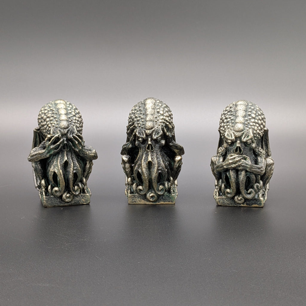 Three small metallic fantasy figurines resembling cthulhu, arranged side by side on a dark grey surface against a gradient grey background.
