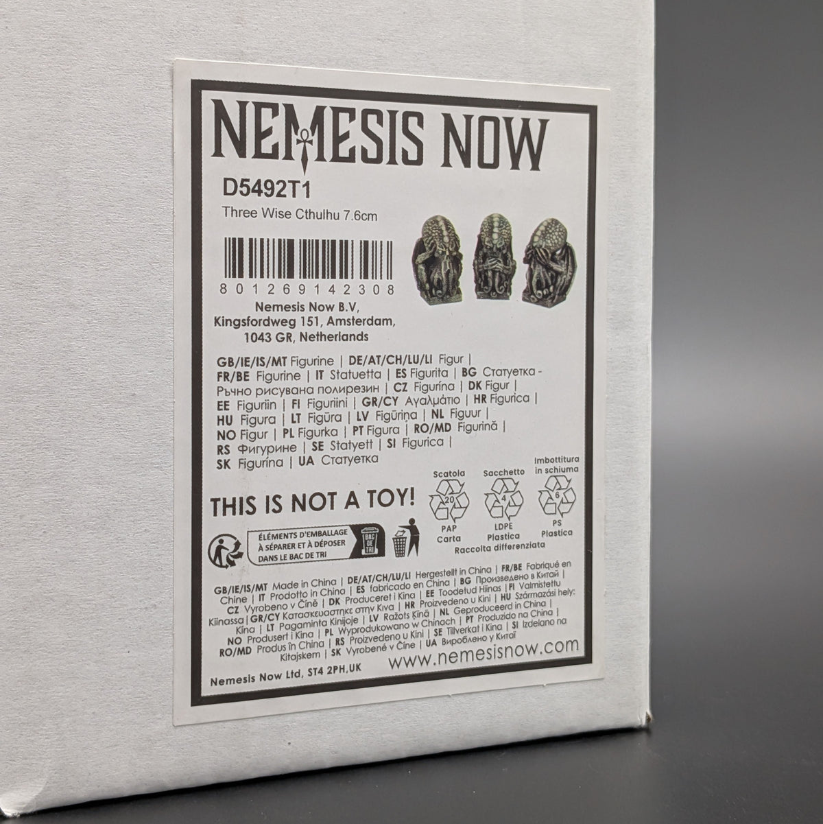 A close-up of a white box with a large printed product label reading 'NEMESIS NOW' and product details for Three Wise Cthulhu, photographed against a dark grey background.