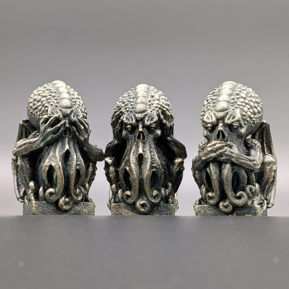 Three gothic Cthulhu figurines with an antique bronze wethered finish, arranged side-by-side on a dark gray surface against a dark grey background.