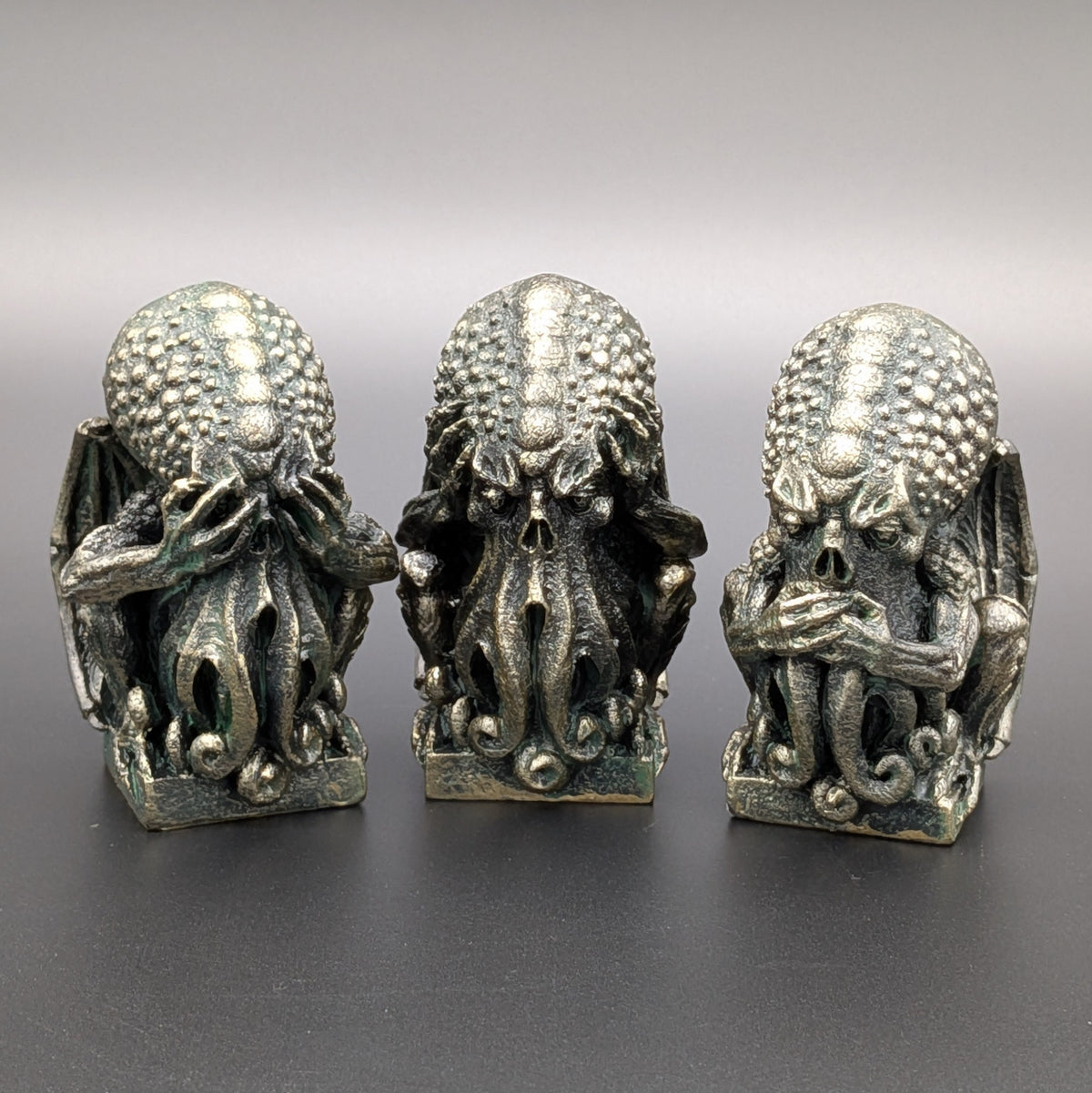 Three small polyresin Cthulhu figurines with a weathered stone patina, arranged side by side depicting "See no evil, Hear no evil, Speak no evil" on a dark grey background.