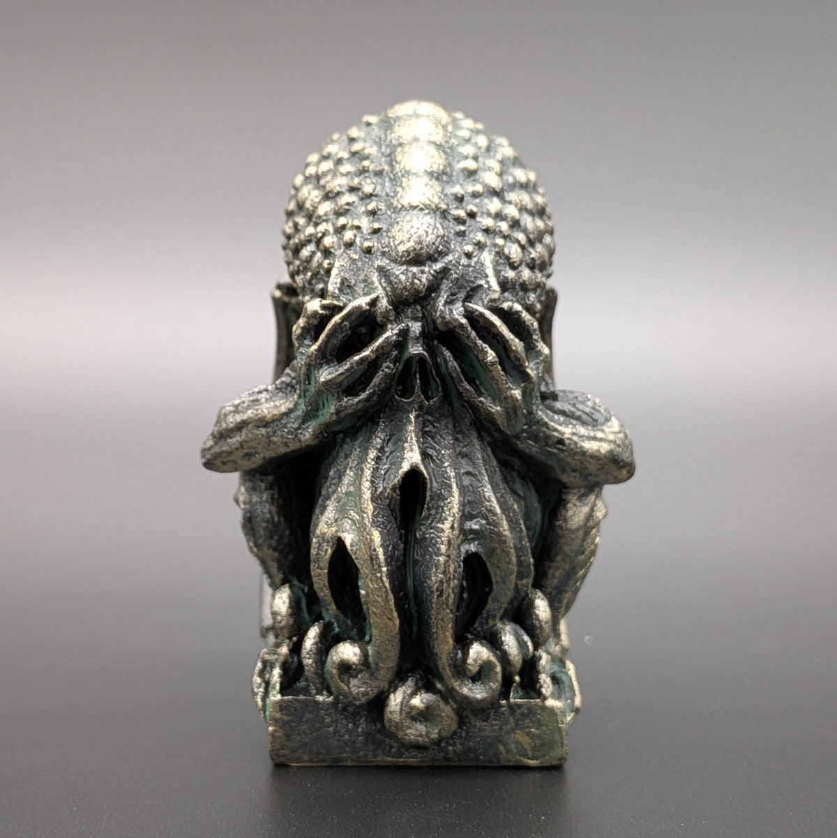 A polyresin Cthulhu figurine with a weathered stone patina & textured skin, depicting "See no evil" by covering his eyes, displayed on a dark grey background.