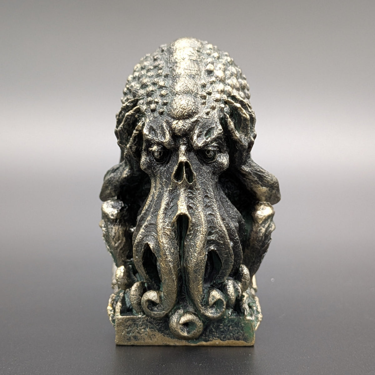 A polyresin Cthulhu figurine with a weathered stone patina & textured skin, depicting "Hear no evil" by covering his ears, displayed on a dark grey background.